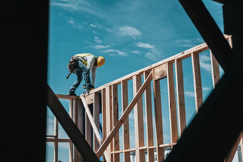 A man in a yellow hard hat and high-vis jacket is standing on a wooden frame of a house, working.